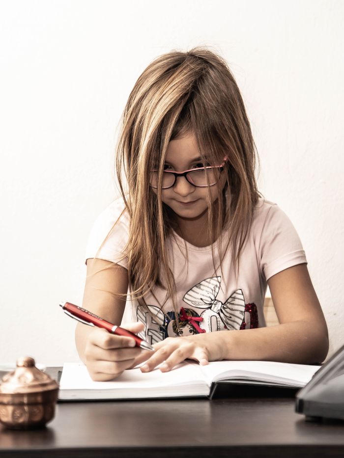 services-01 A young girl with glasses diligently writes in a notebook at a desk, concentrating on her task.