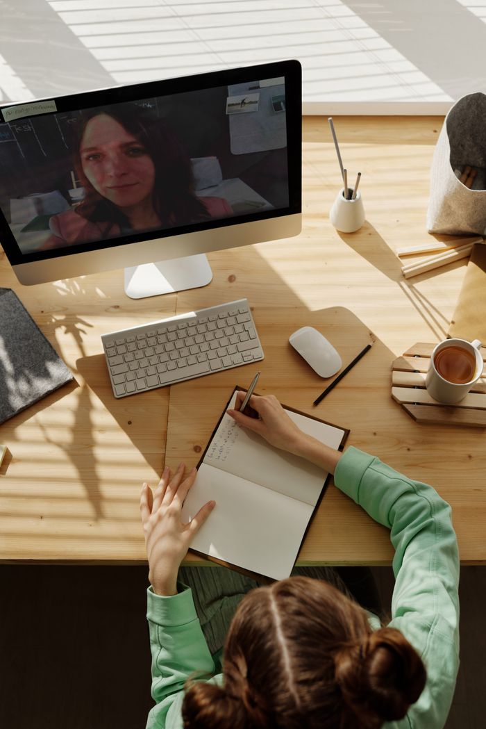 who-we-are A teenager attends an online class from home, taking notes on a notebook during a video call.