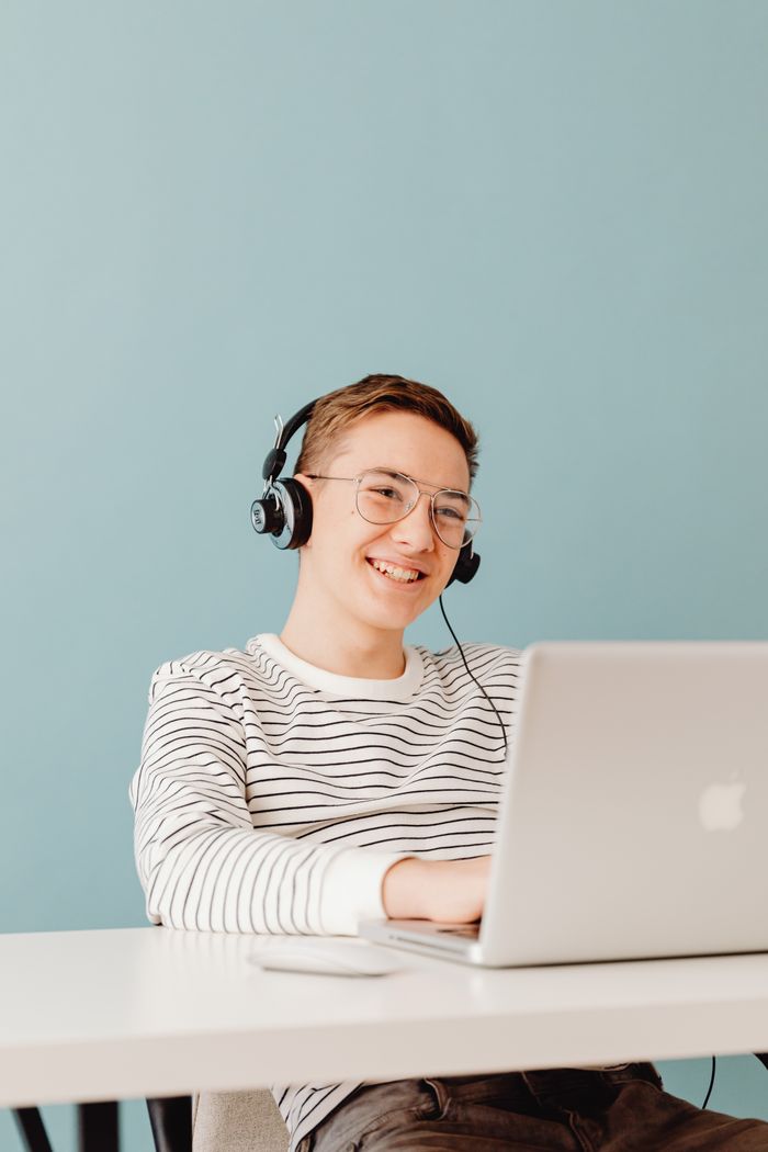 services-03 Young man smiling while using laptop with headphones on, against blue background.